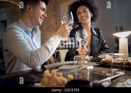 Felice coppia interracial divertirsi parlando, mangiare e bere al ristorante fusion food. Giovani che datano i wineglasses clinking. Foto Stock