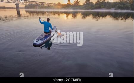 Paddleboard. Uomo seduto su una tavola SUP in mezzo a un lago e godendo dell'alba dorata e della nebbia Foto Stock
