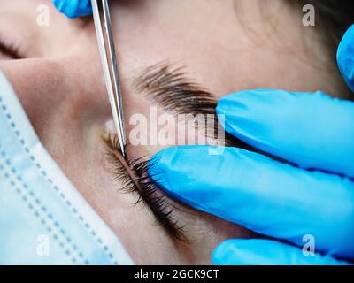 Closeup di raccolto anonimo estetista in guanti di lattice utilizzando pinzette mentre si applicano le ciglia lunghe sulla palpebra della cliente femminile in salone Foto Stock