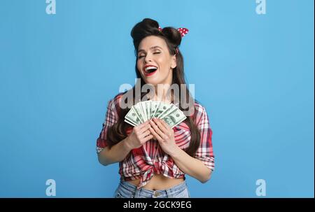 La giovane donna di pinup overjoyed che tiene la somma grande dei soldi vicino a cuore, eccitata sopra la lotteria di vincita su sfondo blu Foto Stock