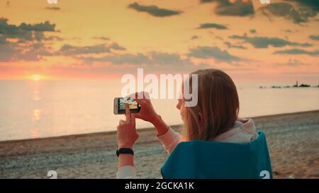 Giovane donna che fa il video del tramonto con smartphone seduto in riva al mare. Vista posteriore Foto Stock