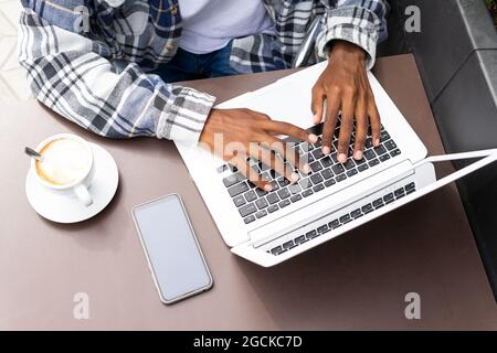 Dall'alto troncato irriconoscibile African American maschio freelancer navigare e lavorare a distanza sul computer portatile in un caffè all'aperto mentre si siede a tavola wit Foto Stock