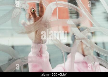 Attraverso un bicchiere di irriconoscibile ragazzo etnico in felpa con cappuccio rosa con brillante pittura creativa manicure sulla finestra trasparente con le dita Foto Stock