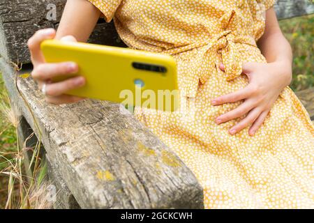 Donna incinta tagliata e irriconoscibile in un abito che tocca la pancia e che si fa autoritratto sul telefono cellulare mentre si siede sulla panchina in campagna in sume Foto Stock