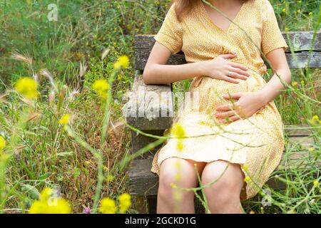 Crop anonima donna incinta in estate vestito a contatto con pancia seduta su panchina in campagna in estate Foto Stock