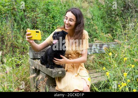 Donna allegra che si scatta da solo con un puppy nero morbido mentre si usa lo smartphone e si siede su una panca di legno in campagna Foto Stock