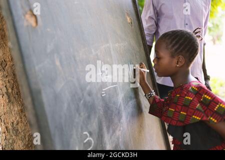 In questa immagine, una piccola ragazza nera concentrata sta scrivendo i numeri su una lavagna durante le classi all'aperto in una comunità rurale nell'Africa occidentale Foto Stock