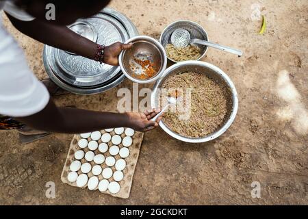 In questa immagine, una cuoca africana nera aggiunge spezie ad un piatto semplice che sta preparando per la sua famiglia, composto solo da cereali, uova bollite e onio fritto Foto Stock