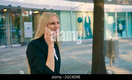 Adulto bionda imprenditrice in tuta seduta sul banco e agitando con la mano la saluto collega sulla strada Foto Stock