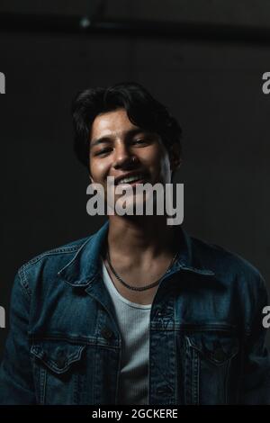 Ritratto di un giovane latino sorridente che guarda la telecamera sotto una luce drammatica e uno sfondo scuro Foto Stock