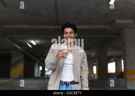 Giovane uomo latino sorridente in abiti casual utilizzando il telefono mentre si trova in piedi nel parcheggio Foto Stock