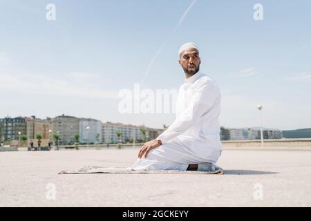 Maschio islamico pieno in abiti bianchi tradizionali in piedi sul tappeto e pregare contro il cielo blu Foto Stock