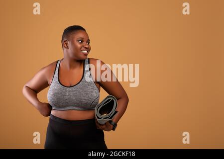 Pronto per la formazione. Ritratto di donna chubby afroamericana che va in palestra, tenendo e trasportando il tappeto arrotolabile in piedi isolato sopra lo sfondo marrone dello studio Foto Stock