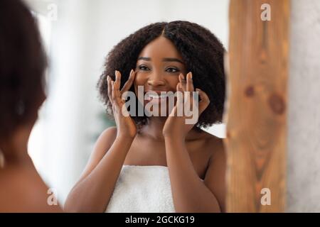 Affascinante signora nera sorridente a se stessa nello specchio, controllando le prime rughe, ammirando la morbida pelle setosa dopo crema viso a casa. Donna afroamericana l Foto Stock