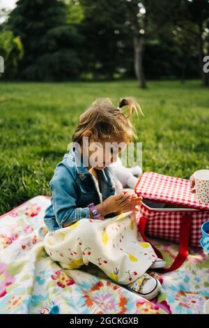 Vista laterale della carina bambina in abito e giacca in denim che gioca su colorata coperta da picnic sul prato verde nel parco estivo Foto Stock