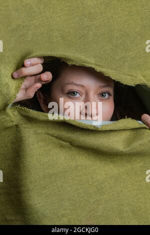 Giovane donna con occhi verdi guardando la macchina fotografica mentre si nasconde dietro un panno verde strappato Foto Stock