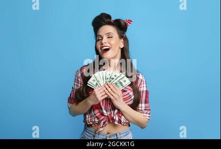 La giovane donna di pinup che tiene la grande somma di denaro vicino a cuore, eccitata oltre la lotteria vincente su sfondo blu studio. Signora millenaria in retrò Foto Stock