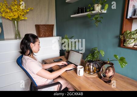 Vista laterale della donna asiatica freelance che legge documenti sul computer portatile mentre si siede al tavolo durante il lavoro remoto Foto Stock