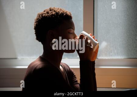 Vista laterale della donna afroamericana con capelli corti bevendo bevande rinfrescanti dalla tazza a casa in mattina Foto Stock