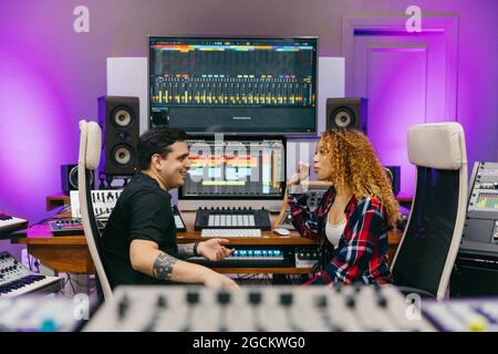 Vista laterale del sound man che parla con una allegra compagna etnica mentre si guarda l'un l'altro tra le attrezzature professionali in studio di registrazione Foto Stock