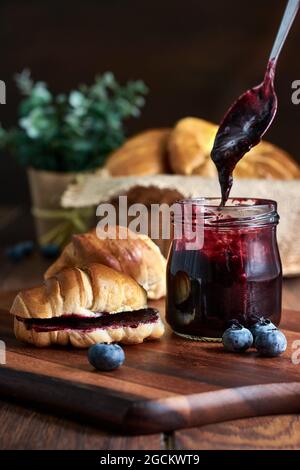 Preparazione di una colazione di croissant con marmellata di mirtilli su un tavolo di legno Foto Stock