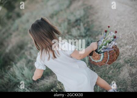 Una bambina in un abito bianco con un cesto di vimini con fiori selvatici. Foto Stock