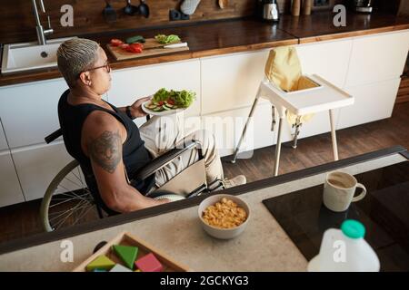 Ritratto ad alto angolo di donna tatuata contemporanea con disabilità in cucina, baby chair in shot, spazio copia Foto Stock