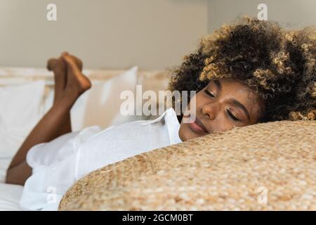 Affascinante donna afroamericana con capelli ricci distesi sul letto con occhi chiusi mentre si riposa a casa Foto Stock