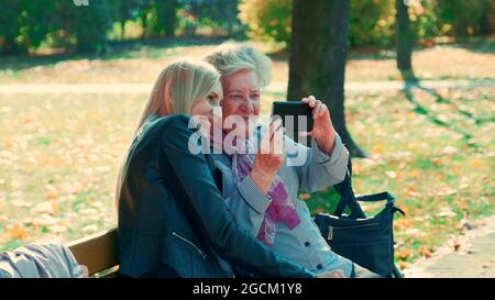 Zoom shot di giovani e vecchie donne che fanno selfie su smartphone in autunno tempo in un bellissimo parco autunno. Si siedono su panchina e sorridono. Foto Stock