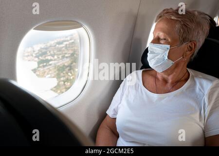 Una donna anziana viaggia. Volare in un aereo che indossa una maschera. Passeggero. Guarda il oblò. Osservare il terreno dall'alto. Stile di vita attivo. Foto Stock