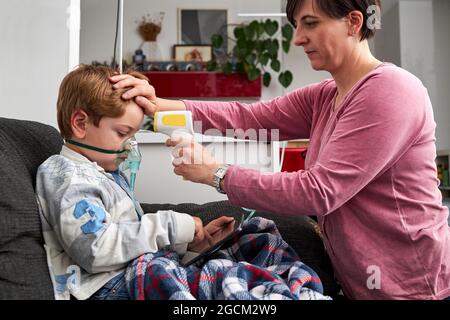 Madre amorevole con termometro a infrarossi misurazione della temperatura del ragazzo che respira in maschera di ossigeno durante la procedura di inalazione e guardare cartoni animati sulla tavola Foto Stock