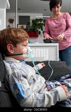 Alto angolo di madre di raccolto che usa il nebulizzatore per l'inalazione di ragazzo seduto in maschera di ossigeno sul divano Foto Stock