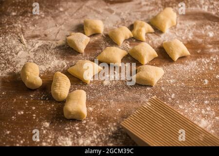 Vista dall'alto di pezzi di pasta morbida e cruda posti su un tavolo di legno coperto di farina vicino al bordo di costoletta durante la preparazione degli gnocchi in cucina Foto Stock