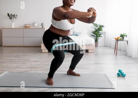 Vista ritagliata di una donna nera curvy squatting con fascia di gomma su allenamento domestico Foto Stock