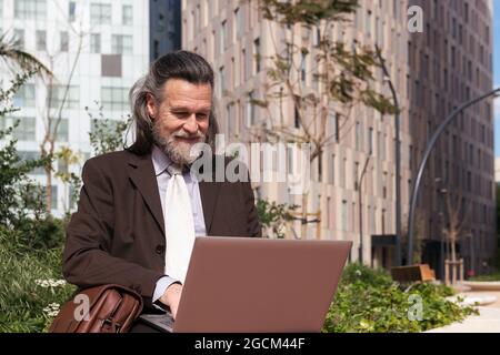 Felice successo grigio capelli bearded maschio in abito elegante utilizzando il computer portatile mentre si siede in strada urbana Foto Stock