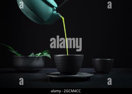 Il sano tè giapponese matcha viene versato dalla teiera verde nella ciotola nera in ceramica durante la cerimonia del tè su sfondo nero Foto Stock