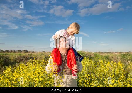 figlio si siede sulle spalle di papà. Entrambi indossano camicie nazionali ricamate ucraine sullo sfondo di un campo di colza in fiore. Educazione patriottica. P. Foto Stock