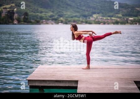 Vista laterale del pacifico equilibrio femminile sulla gamba a Tuladandasana mentre si pratica yoga sul molo di legno vicino al laghetto che guarda via Foto Stock