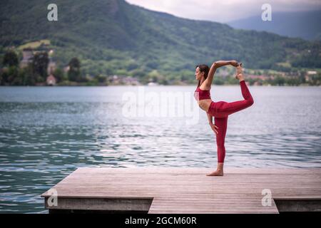 Vista laterale del pacifico equilibrio femminile sulla gamba in Natarajasana mentre si pratica yoga sul molo di legno vicino al laghetto che guarda via Foto Stock