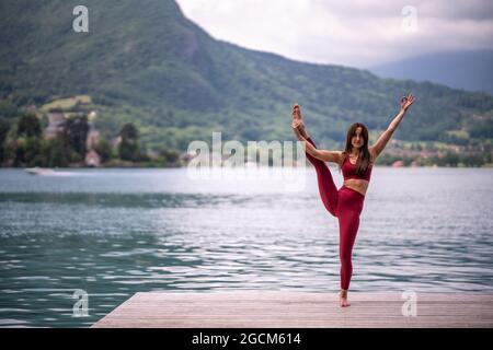 Equilibrio femminile pacifico sulla gamba in Trivikramasana mentre si pratica yoga sul molo di legno vicino al laghetto guardando la macchina fotografica Foto Stock