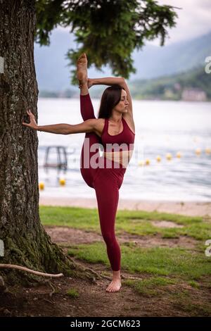 Donna sottile in piedi in Svarga Dvijasana mentre si fa yoga sulla riva dello stagno e appoggiarsi sull'albero Foto Stock