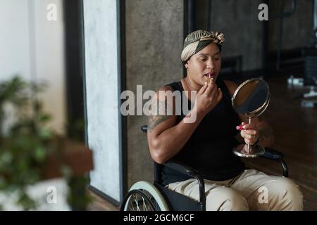 Ritratto della donna afroamericana in sedia a rotelle che si mette sul rossetto mentre tiene lo specchio nell'interno domestico Foto Stock