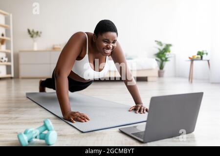 In più, la donna afro di dimensioni in piedi in posizione a tavola o facendo push up vicino al laptop, allenandosi a casa per il tutorial sportivo online Foto Stock