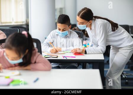 Insegnante femminile in maschera chirurgica viso aiutare piccolo scolaro Foto Stock