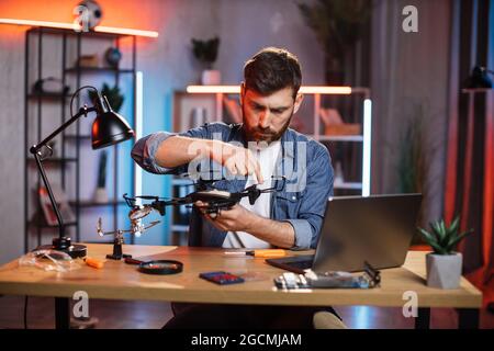 Uomo bearded concentrato usando vari attrezzi per riparare il quadricottero moderno a casa. Ragazzo caucasico in camicia di denim che fissa il drone volante da solo. Foto Stock