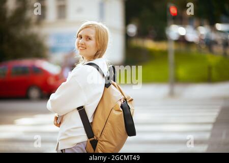 felice ragazza elegante in felpa bianca con zaino che attraversa la passerella e va a scuola all'aperto in città. Foto Stock