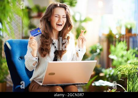 Green Home. Sorridente elegante casalinga di mezza età con lunghi capelli ondulati con carta di credito utilizzando il computer portatile mentre si siede in una poltrona blu nel moderno hou Foto Stock