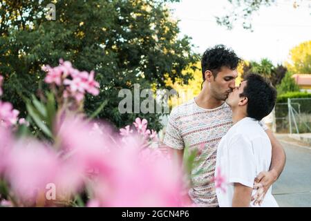 Giovane coppia gay in amore baciando in natura con fiori in primo piano. Due giovani amanti all'aperto in estate su una strada. Felice coppia caucasica innamorata Foto Stock