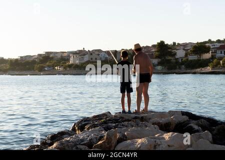 Rogoznica, Croazia-8 luglio 2021: Nonno insegna a suo nipote trucchi di pesca sulla costa rocciosa di Rogoznica, Croazia al tramonto Foto Stock