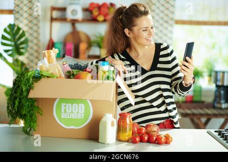 Consegna di cibo. Donna giovane felice con scatola di cibo in cucina utilizzando applicazioni smartphone. Foto Stock
